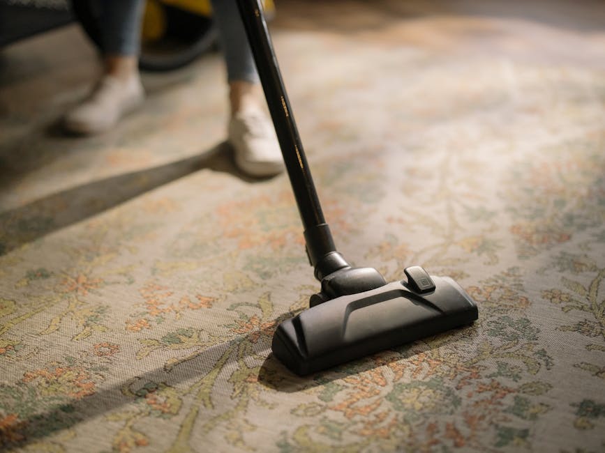 A close-up view of an upright vacuum cleaner with a transparent dust container filled with dirt and debris, connected to a flexible, patterned hose that leads to a floor brush attachment on a pink car