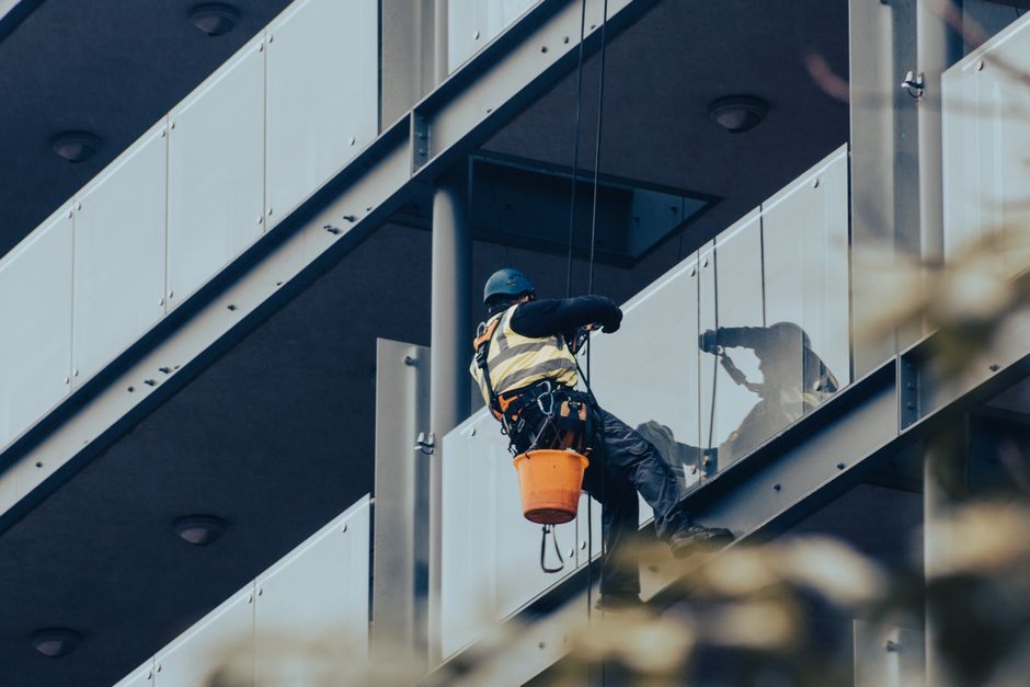 A professional window cleaner wearing a safety helmet, harness, and reflective vest is performing surface cleaning on the glass panels of a balcony at a modern residential building. The worker is usin