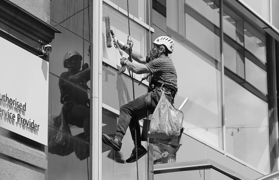 A professional window cleaner wearing a safety helmet, harness, and reflective vest is performing surface cleaning on the glass panels of a balcony at a modern residential building. The worker is usin