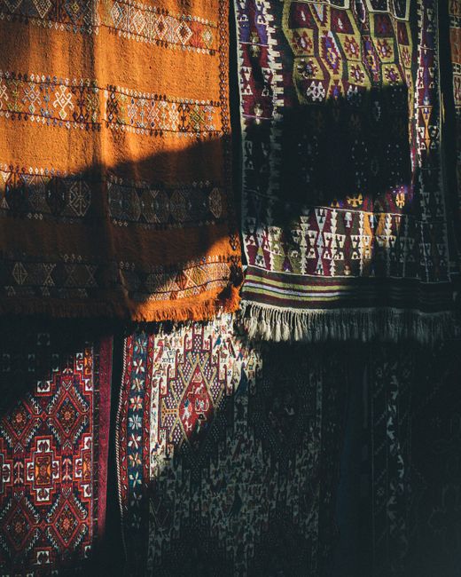 A display of multiple colorful, patterned rugs hanging from wooden hooks on a wooden beam inside a shop near Fulham. The rugs feature intricate designs with floral and geometric motifs in hues of red,