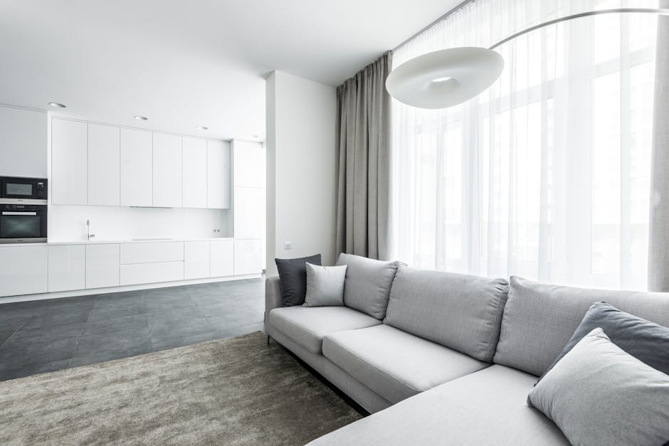 A bright, modern living room with a light gray fabric sofa adorned with a mix of dark and light gray cushions, positioned near large floor-to-ceiling windows covered with sheer white curtains and heavier gray drapes. The room features a white ceiling with recessed lighting and a contemporary white pendant light fixture. The flooring transitions from dark gray tiles in the kitchen area to a soft, beige carpet in the living space, which appears clean and well-maintained. The kitchen area displays sleek white cabinets with minimal handles, integrated appliances, and a stainless steel sink, all arranged along one wall, contributing to a tidy and hygienic appearance. Natural daylight streams through the sheer curtains, illuminating the space evenly. This setup exemplifies domestic surface cleaning and maintenance practices, aligning with the focus of Fulham Carpet Cleaners on upholstery and surface care in period homes, as discussed in the 'Parsons Green upholstery cleaning tips for period homes, Fulham' page.